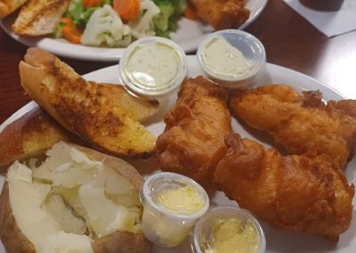 Fish and chips meal with golden battered fish, crispy potato wedges, baked potato, and dipping sauces on white plates