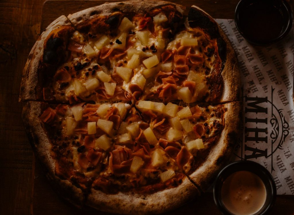 A close-up shot of a Hawaiian pizza with ham and pineapple slices on a rustic wooden table.