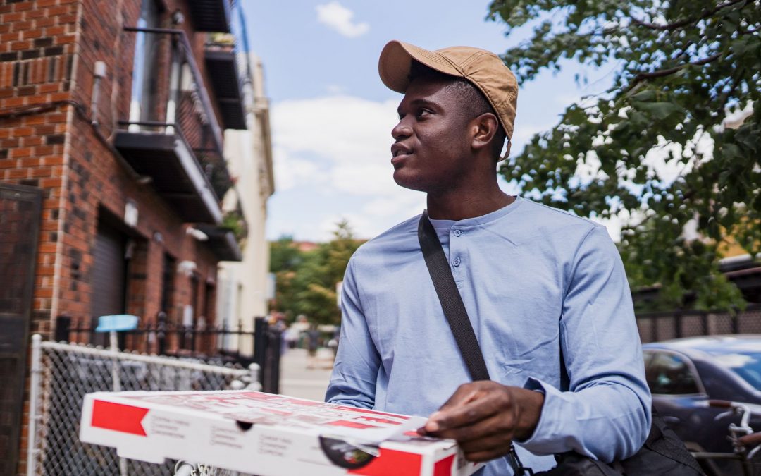 A delivery person with a pizza box walking outside on a sunny day, wearing a cap and blue shirt.