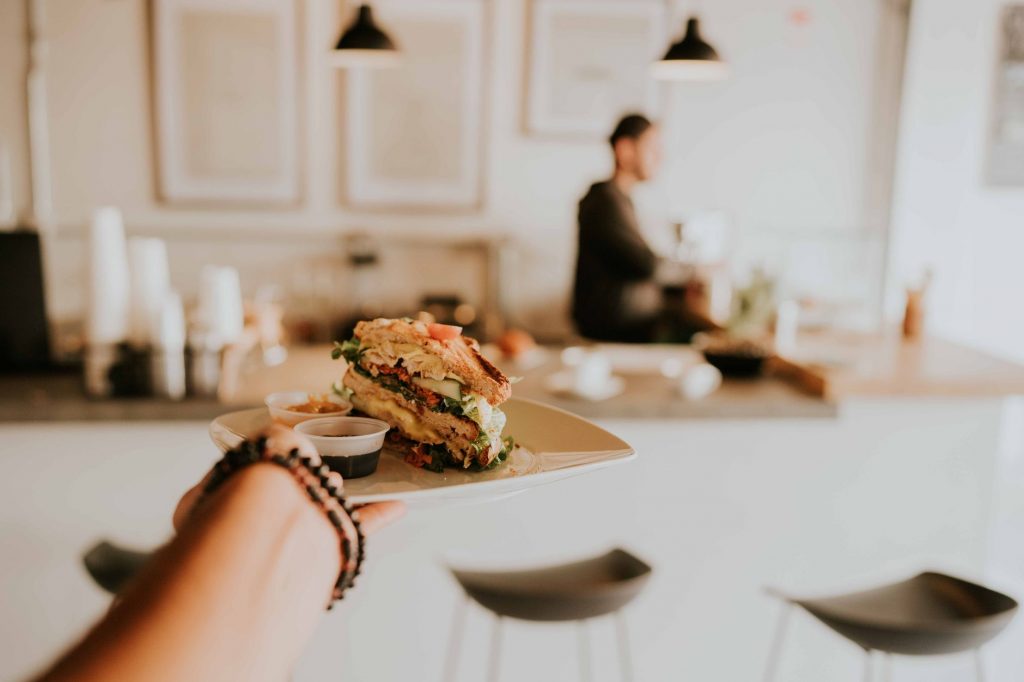 A hand holds a gourmet sandwich on a plate in a cozy café with a blurred barista in the background.