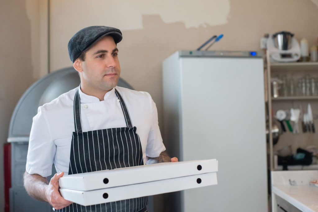 Chef wearing apron and flat cap holding pizza boxes in a kitchen setting.