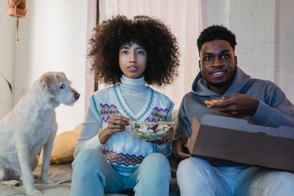Concentrated young African American couple with curly hairs in casual outfits eating takeaway salad and pizza while watchi…