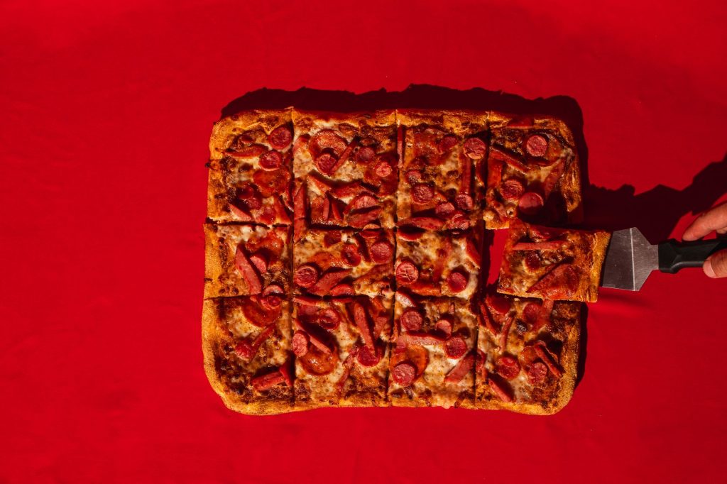 Rectangular pepperoni pizza being served on a vibrant red surface.