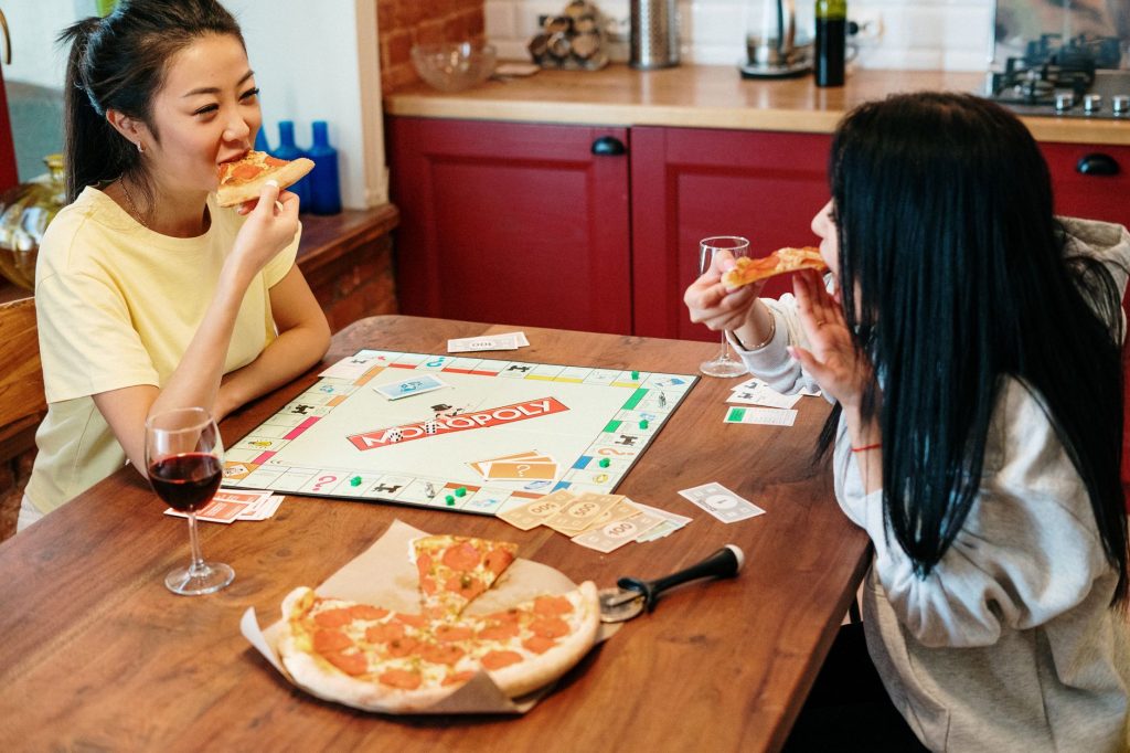 Two friends enjoying a casual game of Monopoly with pizza and wine indoors.
