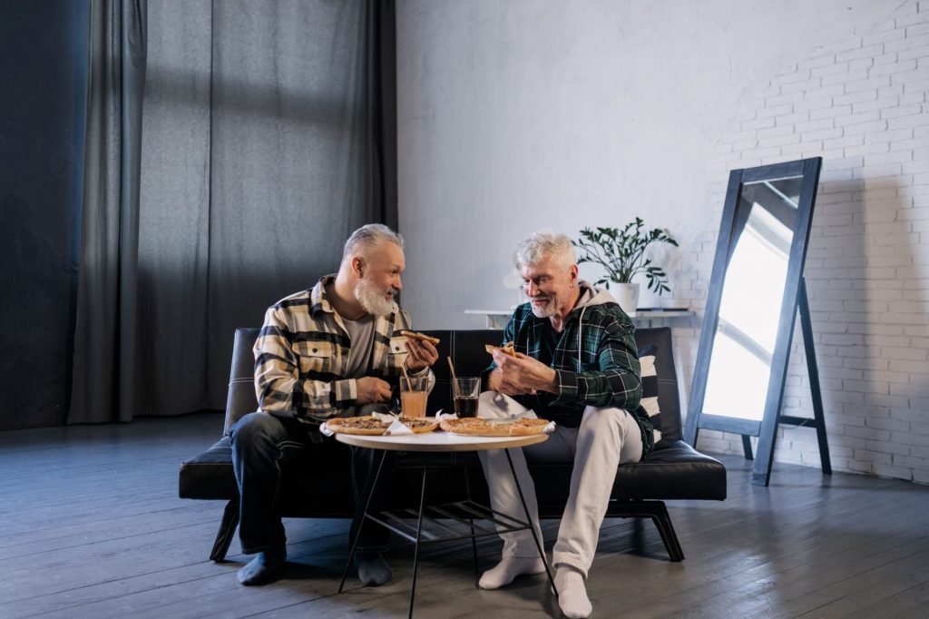 Two senior men enjoying pizza and drinks at a table inside a modern living room.