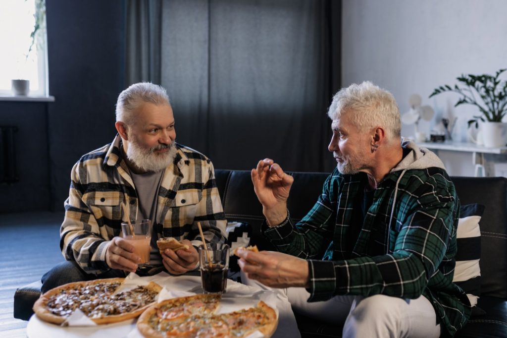 Two senior men enjoying pizza and drinks while chatting in a cozy indoor setting.