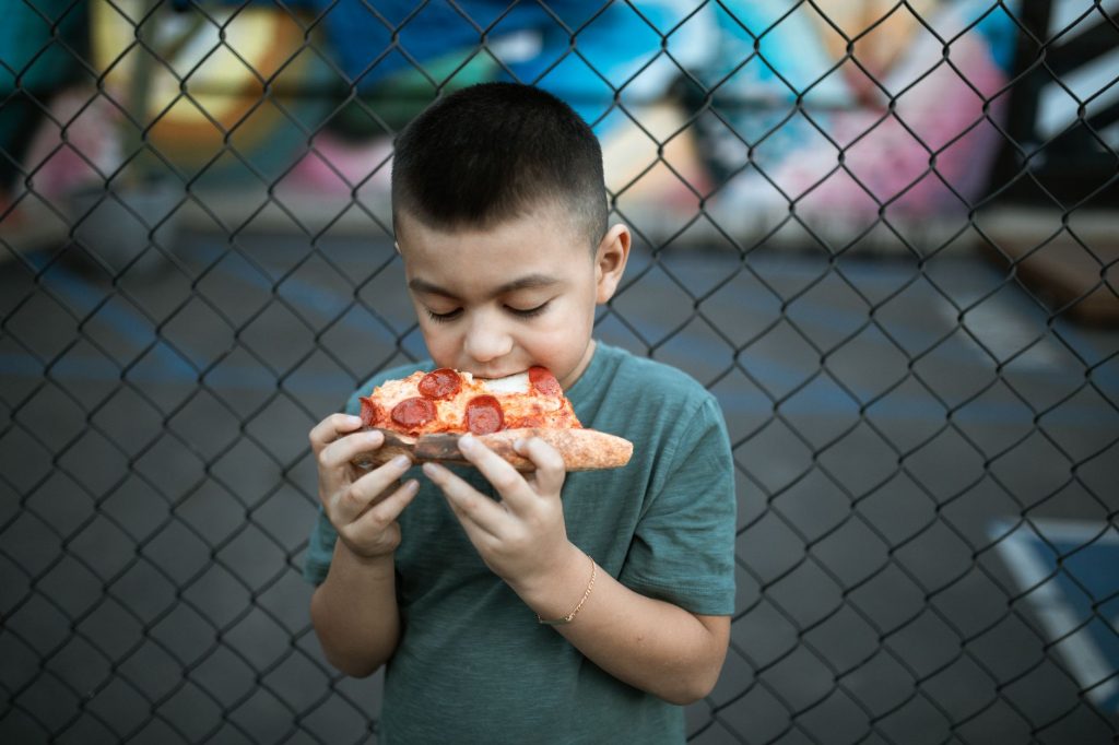 Young boy savoring a slice of pepperoni pizza against a chain-link fence backdrop.